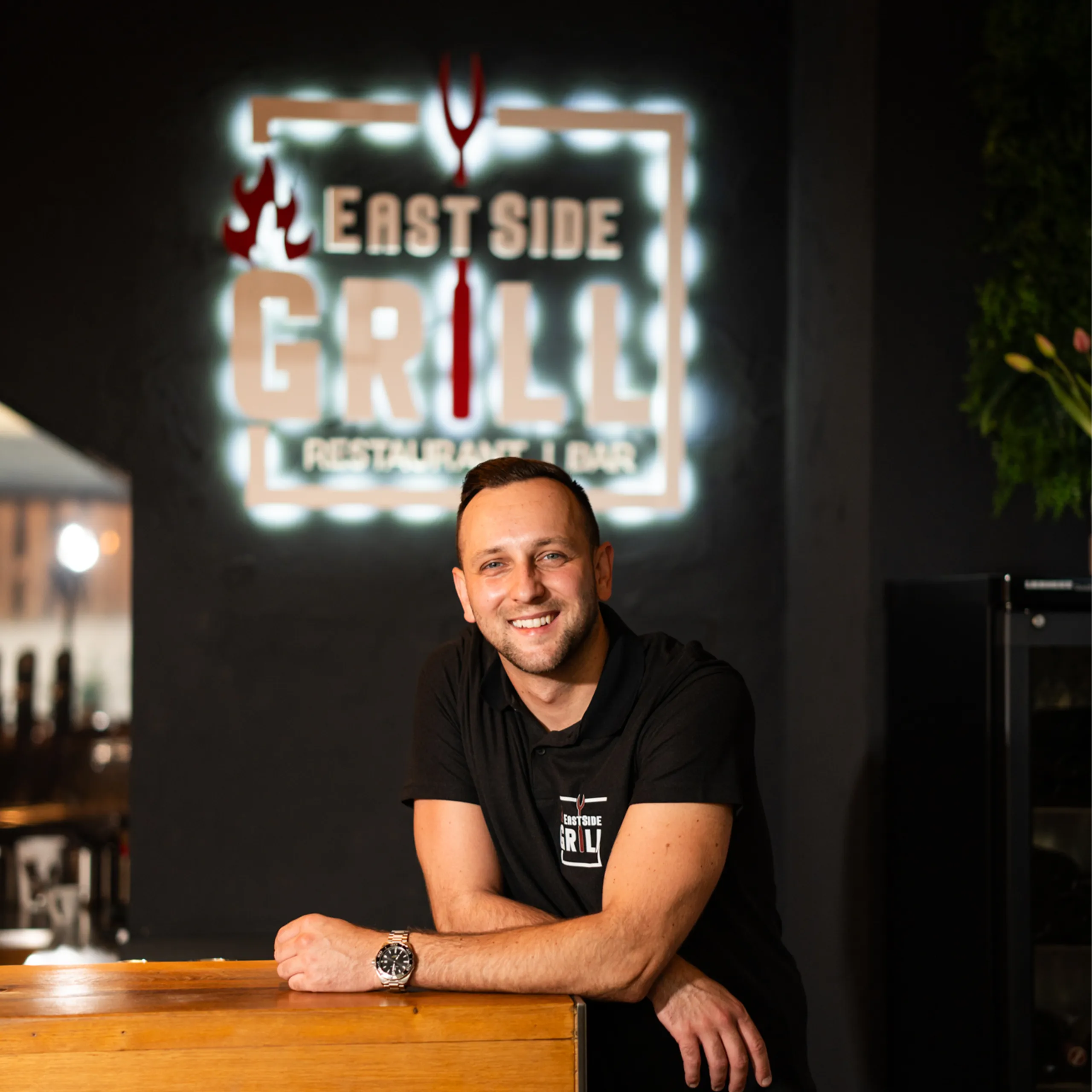 Zeljko, zweiter Inhaber des EastSide Grill, im Restaurant vor dem beleuchteten Logo